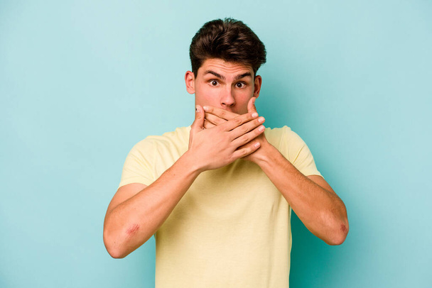 Junger kaukasischer Mann isoliert auf blauem Hintergrund schockiert Mund mit Händen bedecken. - Foto, Bild