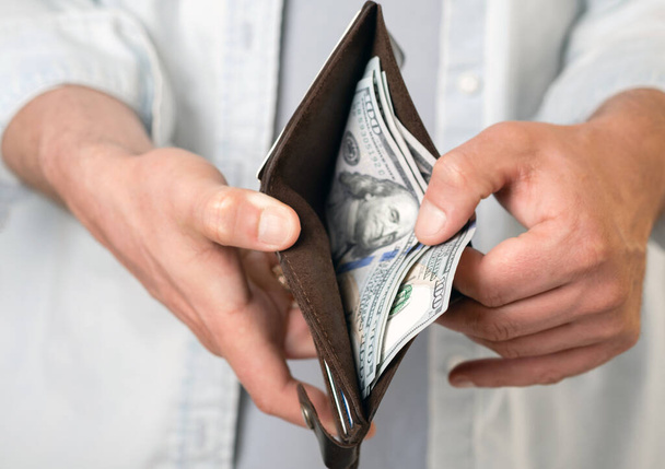 Muž v neformálních šatech drží mužskou hnědou peněženku s penězi a stodolarovými bankovkami v rukou. Modrá košile. - Fotografie, Obrázek