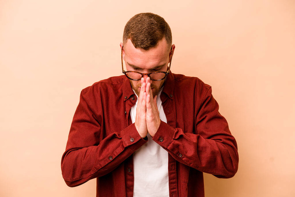 Молодий кавказький чоловік ізольований на бежевому фоні молячись, демонструючи відданість, релігійна людина шукає божественне натхнення
. - Фото, зображення