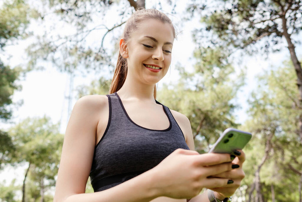 Cheerful brunette woman wearing sportive clothes on city park, outdoors looking at the phone screen and using phone. Messaging with friends, watching video or scrolling on social media. - Photo, image