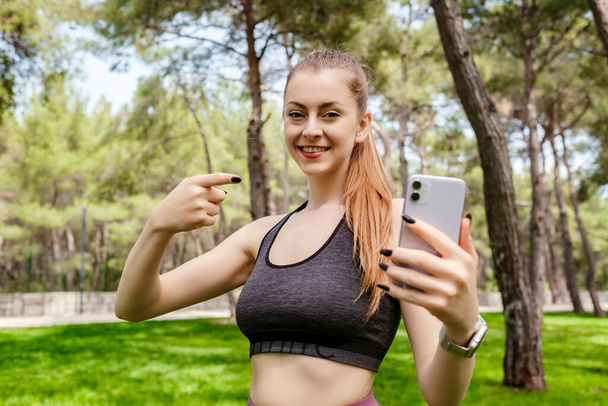 Femme brune joyeuse portant des vêtements de sport sur le parc de la ville, à l'extérieur tenant un téléphone et pointant du doigt le téléphone avec le visage souriant. Elle regarde la caméra.. - Photo, image