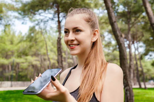 Happy sportive woman wearing sports bra standing on city park, outdoors using her mobile on speaker phone holding the device in front of face. Happy sportive woman talking with friends on phone. - Photo, Image