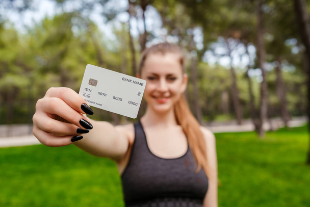 Young woman smiling confident wearing sports bra standing on city park, outdoors showing plastic credit or debit card while holding mobile phone. Online shopping, banking and transaction concepts. - Photo, Image