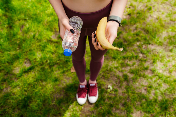 Şehir parkında elinde muz ve su şişesiyle duran spor sutyen giyen güzel esmer kadın. Açık hava sporu ve sağlıklı yaşam kavramları. - Fotoğraf, Görsel