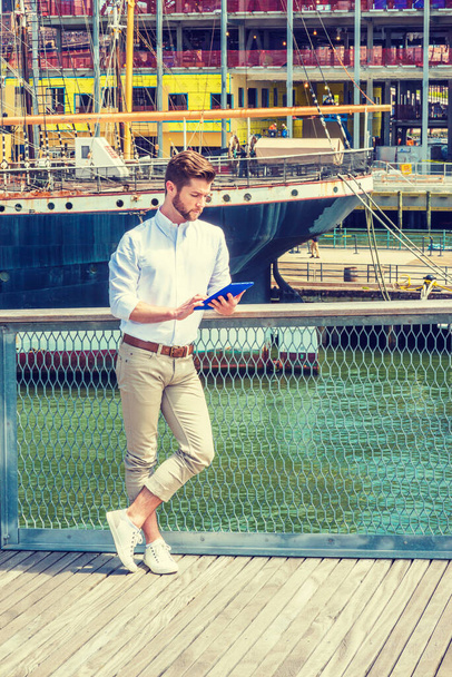 Modern Reading. American Man with beard traveling in New York, wearing white shirt, tan pants, sneakers, standing on deck, reading blue tablet computer. Boat on background.  - Photo, Image