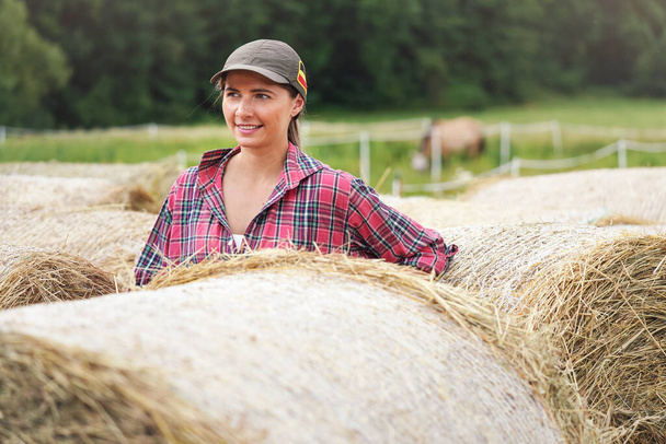 Young woman in shirt and cap smiling, hay stack rolls near her - Foto, imagen