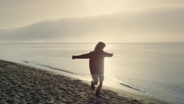 Opgewonden vrouw die zich omdraait op het strand. Gelukkig meisje dansend aan zee. Vrouwelijke hipster springt aan de oceaan. Stijlvolle vrouw die handen opsteekt. Jongedame die zich buiten gelukkig voelt. Uitzicht op zee bij zonsopgang - Video