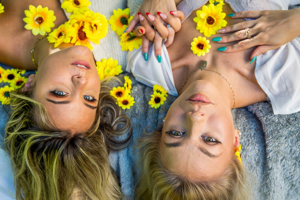 Dwie piękne blondynki modelki poza domem z dzikimi kwiatami ciesząc się wiosenną pogodą - Zdjęcie, obraz