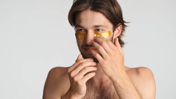 Красива брюнетка бородатий чоловік наносить золоті плями очей, піклуючись про свою шкіру на білому тлі. Концепція краси
 - Фото, зображення