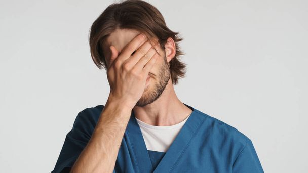 Zmęczony lekarz trzymający rękę na twarzy coś poszło nie tak w pracy w szpitalu. młody stażysta ubrany w mundur patrząc wyczerpany na białym tle - Zdjęcie, obraz