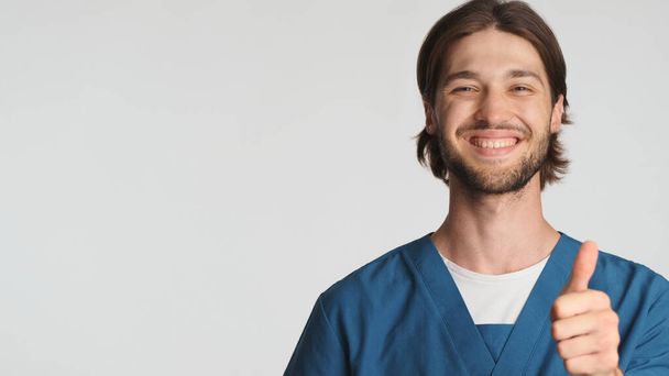 Positive bärtige männliche Arzt lächelnd den Daumen nach oben über weißem Hintergrund. Attraktive Praktikanten in Uniform sehen selbstbewusst aus und zeigen vereinzelt zugelassene Zeichen. Gefällt mir. Kopierraum - Foto, Bild