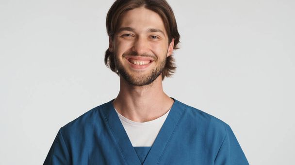 Houkutteleva parrakas mieslääkäri hymyilee kameralle valkoista taustaa vasten. Kaunis harjoittelija pukeutunut univormu näyttää onnelliselta poseeraa studiossa. Sairaanhoitaja  - Valokuva, kuva