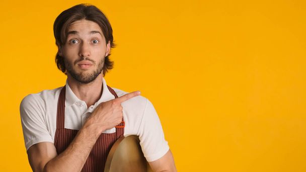 Astonished bearded man wearing apron pointing aside with fingers isolated. Young waiter looking amazed showing copy space over yellow background - Photo, Image
