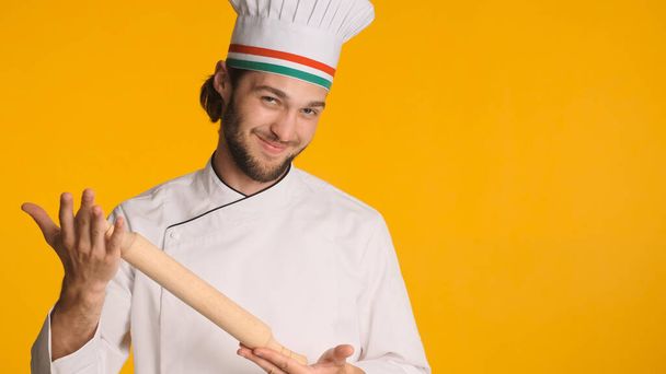 Atraktivní mužský kuchař v bílé uniformě a čepici s dřevěným válečkem v rukou, záludně pózující před kamerou na barevném pozadí. Mladý kuchař připraven udělat dezert  - Fotografie, Obrázek