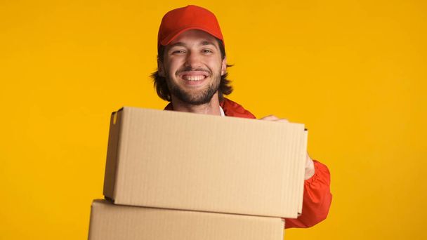 Beautiful delivery man wearing red uniform holding cardboard boxes over colorful background. Male bearded courier enjoying his work standing with parcel boxes in studio - Photo, Image