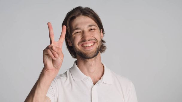 Homme barbu joyeux montrant signe de paix souriant sincèrement à la caméra sur fond gris. Jeune homme positif posant en studio - Photo, image