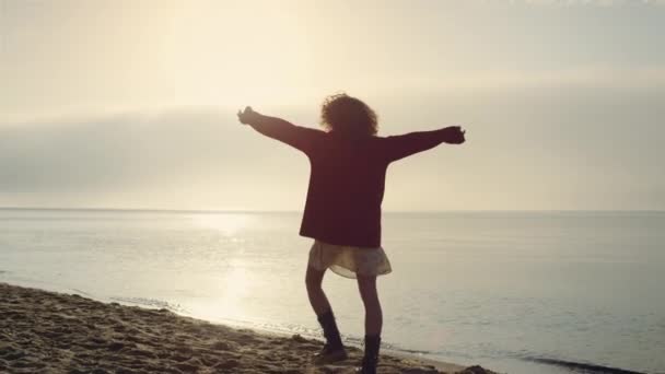 Positive woman raising hands at seaside. Emotional girl felling happy. Silhouette model turning around. Cheerful lady walking on ocean beach at sunrise. Stylish woman enjoying freedom. Victory concept - Footage, Video