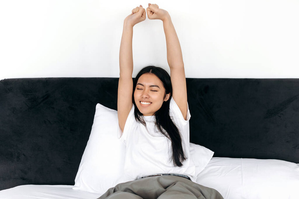 Счастливая азиатская брюнетка просыпается, делает растяжку утром в выходные, сидя на кровати в спальне, поднимает руки с закрытыми глазами, счастливо улыбаясь - Фото, изображение