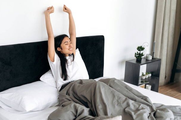 Здорова азіатська брюнетка щаслива дівчина в білій футболці, прокидається з посмішкою і розтягує руки після хорошого сну, сидячи в затишному ліжку, починає свій день з великим настроєм, добре спати
 - Фото, зображення