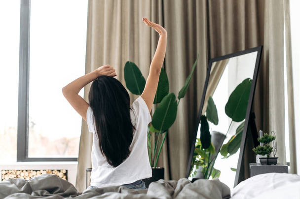 Heureuse fille brune asiatique dans un t-shirt blanc, se réveille et étire les bras après bien dormir assis dans un lit confortable retour à la caméra, commence sa journée avec une bonne humeur, ayant une bonne nuit de sommeil - Photo, image