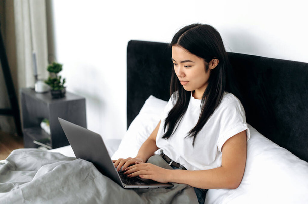 Lovely happy asian brunette girl dressed in a basic white t-shirt, is sitting in a bed in modern bedroom, uses her laptop, chatting with friends in social media, checking email, browsing news, smiling - Foto, imagen
