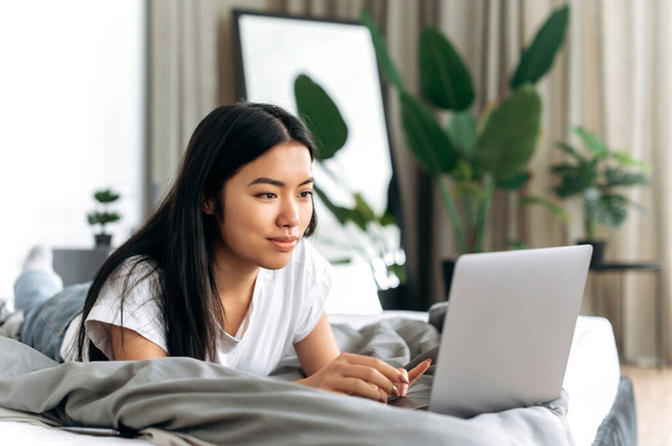 Tecnologia wireless. Positiva ragazza bruna cinese in t-shirt bianca di base, è sdraiata su un letto in una stanza, utilizza il suo computer portatile, chatta con gli amici sui social media, controlla le e-mail, sfoglia le notizie, sorride - Foto, immagini