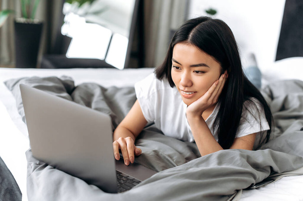 Hezké, pozitivní asijské brunetky dívka v bílém tričku, ležící doma na posteli, používá notebook, prochází sociální sítě, chatuje online s přáteli, čte zprávy, usmívá se, relaxuje doma o víkendu - Fotografie, Obrázek