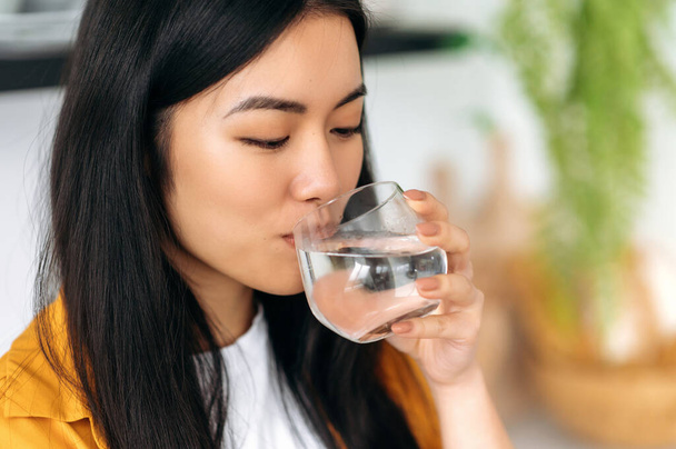 Leading a healthy lifestyle. Close-up of a happy Asian girl, drinks a glass of clean water with pleasure, care her health, balances fluids in the body, replenishes energy and strength in the body - Foto, Bild