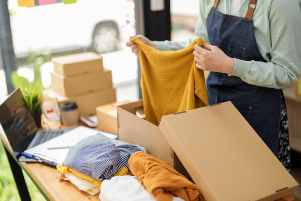 Portrait of a small startup Asian female entrepreneur SME owner picking up a yellow shirt before packing it in an inner box with a customer. Online Business Ideas and Freelance. - Fotografie, Obrázek