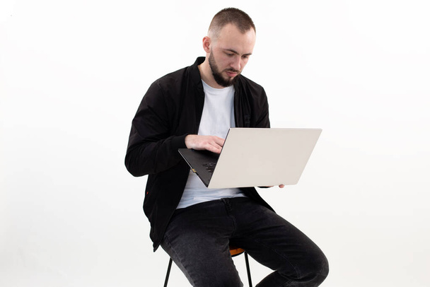 Homme concentré avec barbe à l'aide d'un ordinateur portable sur chaise, travailler en ligne. Succès d'homme d'affaires, studio sur fond blanc. E-commerce et travail à distance à l'intérieur. Gestionnaire sérieux travaillant sur PC, la technologie informatique.  - Photo, image
