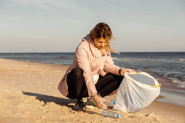 Woman activist and assistant cleaning sandy beach from underwater trash, sea or ocean with plastic waste. Collect bottles to bag. Ecological pollution. Sustainable development. Environmental campaign - Foto, Imagem
