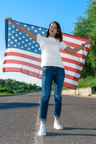 Portret uśmiechniętej młodej kobiety trzymającej amerykańską flagę. USA świętują 4 lipca. Patriotyczna młoda kobieta powiewa amerykańską flagą na pięknym polu.  - Zdjęcie, obraz