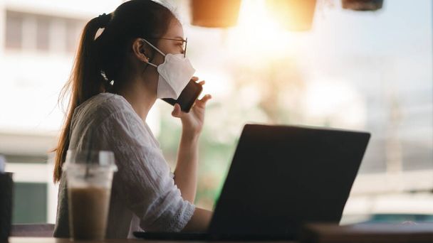 επιχειρηματίας κάθεται στο γραφείο πίσω από το laptop της και μιλάει με κάποιον στο κινητό της ενώ εργάζεται από το σπίτι. - Φωτογραφία, εικόνα
