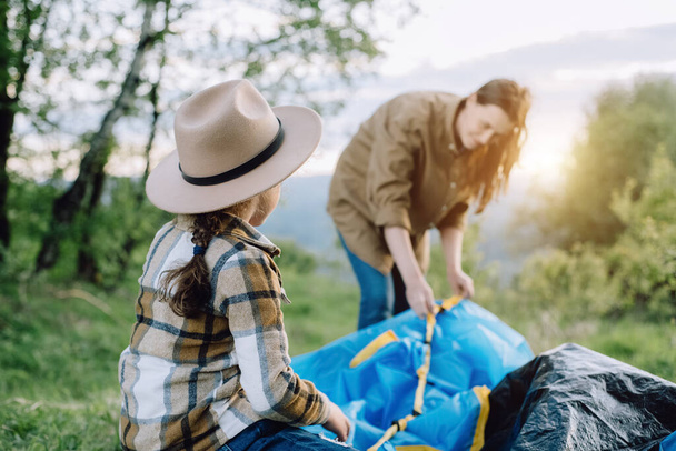 Side view of little daughter in hat and young mother sets up tent near background green forest during sunset or sunrise, outdoor recreation in high mountains. Travel, adventure, wanderlust concept - 写真・画像
