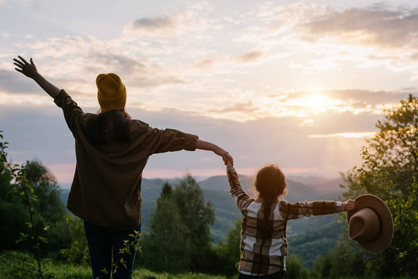 Silhouette of young mother and little daughter kid jumping and waving their hands on background summer mountains during sunset or sunrise. Family rejoicing and having fun. Travel and camping concept - Foto, Bild