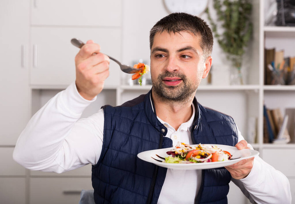 Jeune homme regardant salade de légumes et manger à la table à l'intérieur - Photo, image