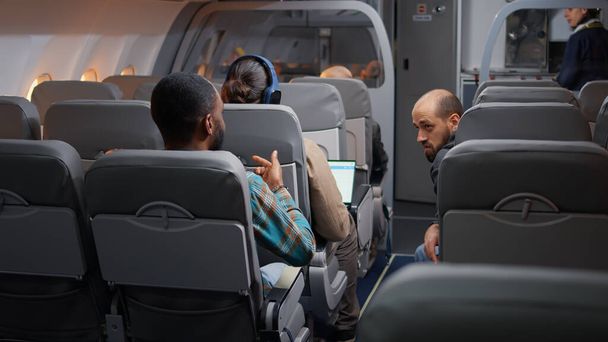 Różnorodni podróżnicy rozmawiający w samolocie przed startem, lecący z międzynarodowymi liniami lotniczymi na wakacje. Podróż samolotem ze stewardessą, zarobkowe drogi oddechowe. - Zdjęcie, obraz