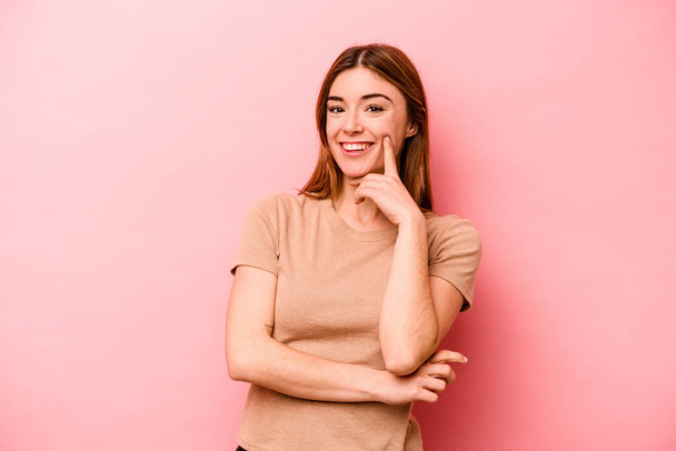Νεαρή καυκάσια γυναίκα απομονωμένη σε ροζ φόντο χαμογελώντας χαρούμενη και με αυτοπεποίθηση, αγγίζοντας το πηγούνι με το χέρι. - Φωτογραφία, εικόνα
