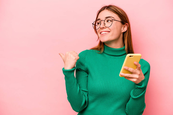 Nuori valkoihoinen nainen pitää matkapuhelinta eristettynä vaaleanpunaisiin taustapisteisiin peukalo sormi poispäin, nauraen ja huolettomana. - Valokuva, kuva