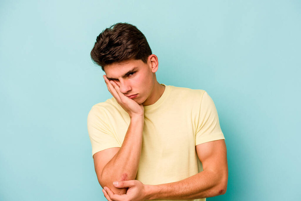 Young caucasian man isolated on blue background who is bored, fatigued and need a relax day. - Foto, afbeelding