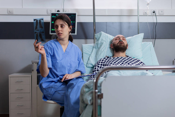Hastaların iyileşme sürecini izleyen hastalara kemik radyografisi gösteren asistana yakın çekim. Hasta adam yatakta dinleniyor. Terapi sırasında hastane koğuşunda hastalık tedavisini tartışıyorlar. - Fotoğraf, Görsel