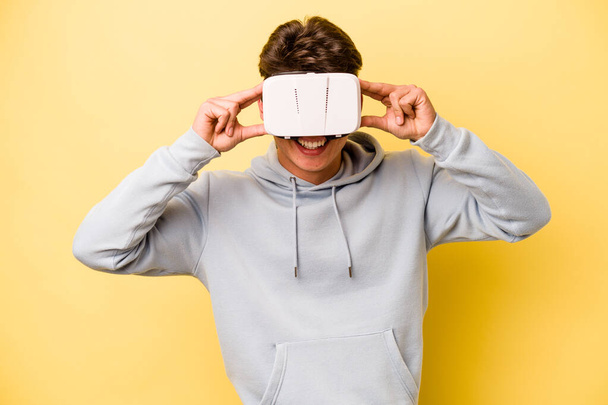 Jeune homme caucasien portant des lunettes virtuelles isolé sur fond jaune - Photo, image