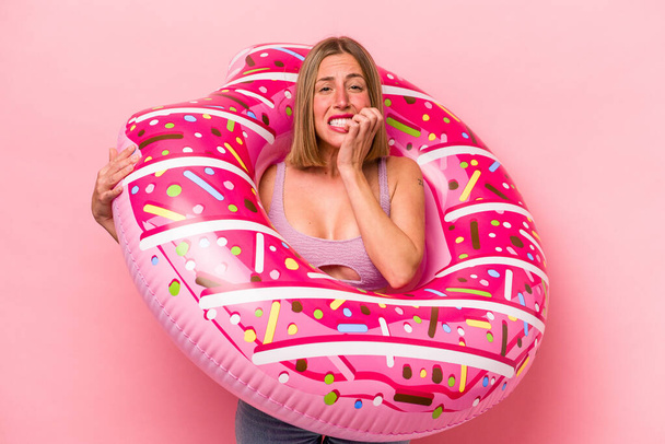 Mujer caucásica joven sosteniendo un colchón de aire aislado sobre fondo rosa mordiendo las uñas, nervioso y muy ansioso. - Foto, imagen