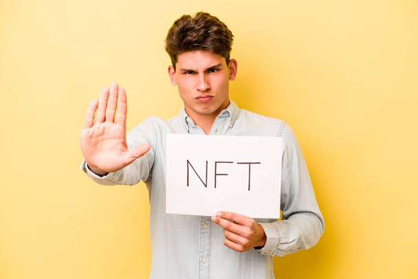 Молодой кавказский мужчина с плакатом NFT на желтом фоне стоит с протянутой рукой, показывая стоп-сигнал, препятствуя вам. - Фото, изображение