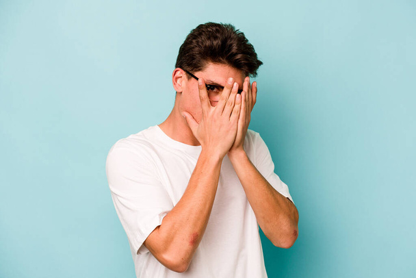 Young caucasian man isolated on blue background blink through fingers frightened and nervous. - Photo, Image