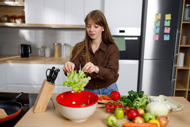 Jonge vrouw die verse salade maakt en sla in de kom gooit. Vrouwelijke veganistische koken gezond voedsel - Foto, afbeelding