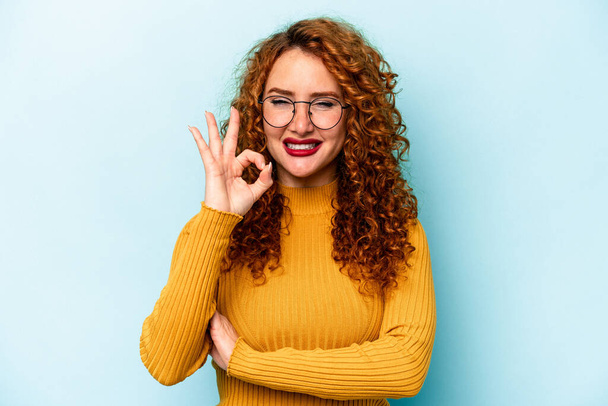 Young ginger caucasian woman isolated on blue background winks an eye and holds an okay gesture with hand. - Fotoğraf, Görsel