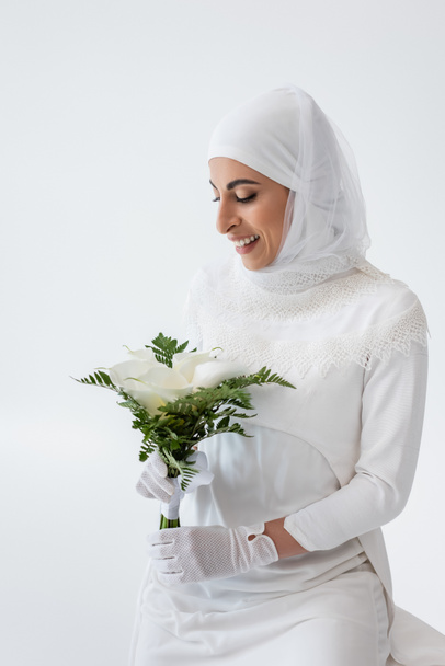 happy muslim bride in gloves and wedding dress holding calla lily flowers isolated on grey - Фото, изображение