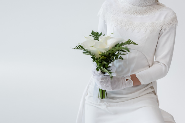 bijgesneden weergave van de bruid in handschoenen en trouwjurk met calla lelie bloemen geïsoleerd op grijs - Foto, afbeelding