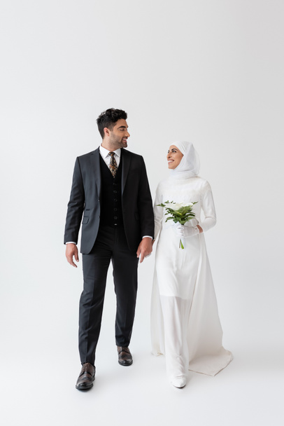 novia musulmana en vestido de novia celebración ramo de lirio de cala y mirando novio feliz en traje en gris - Foto, Imagen